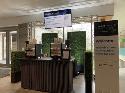 Coffee bar setup at Schuylkill Alumni event, with welcome sign, TV, and green hedge backdrop.