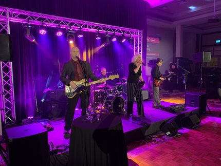 Band performing on stage under purple lights at an event. Musicians play instruments: bass, drums, guitar, and vocals.