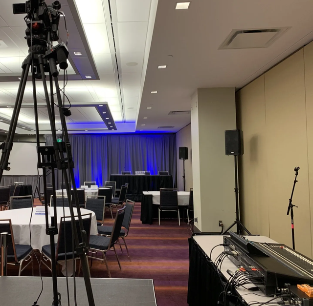 Conference room set up for a presentation. Cameras on tripods, tables with chairs, audio equipment, and a stage with a blue curtain.
