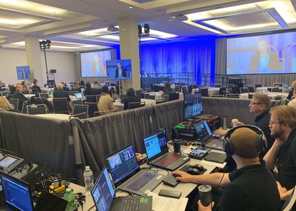A conference room with attendees, multiple screens, and a video production crew working on laptops.