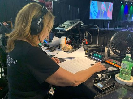 Woman at a soundboard with headphones on, working on a script. A screen and fan are behind her.