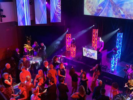People dancing at an event with a DJ on stage, illuminated by colorful lights.