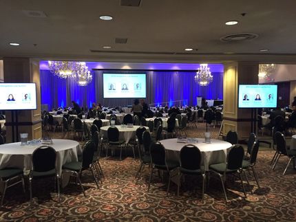 A banquet hall set for a conference with round tables, screens, and a stage with a presentation.