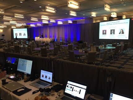 Conference room setup with stage, screens, tables, and laptops in the foreground. Blue and white lighting.