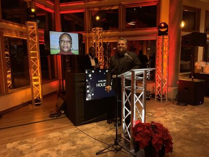 Man speaking at podium, DJ booth with screen displaying a person's image, and red holiday lighting.