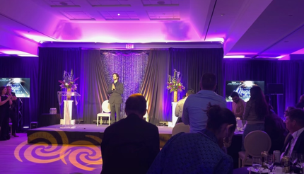 Man speaking on stage at event. Purple lighting, audience seated, black backdrop.