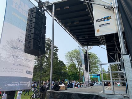Stage setup with stacked speakers, banners, and an audience in a park setting.