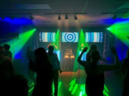People dancing in a club with colorful green and blue stage lights. Central video screens display abstract designs.