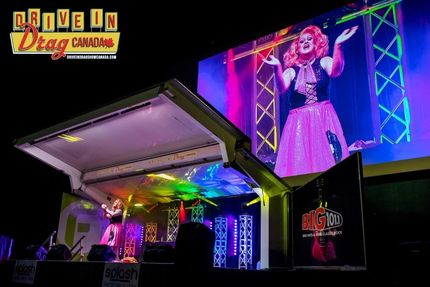 Drag queen on stage at a drive-in show, wearing pink dress and performing under lights.