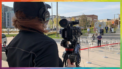 A person filming an outdoor event with a professional camera on a tripod. Empty chairs and ribbon are set up for an event.