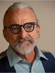 Man with glasses and grey beard, wearing a blue vest and earrings, smiling.