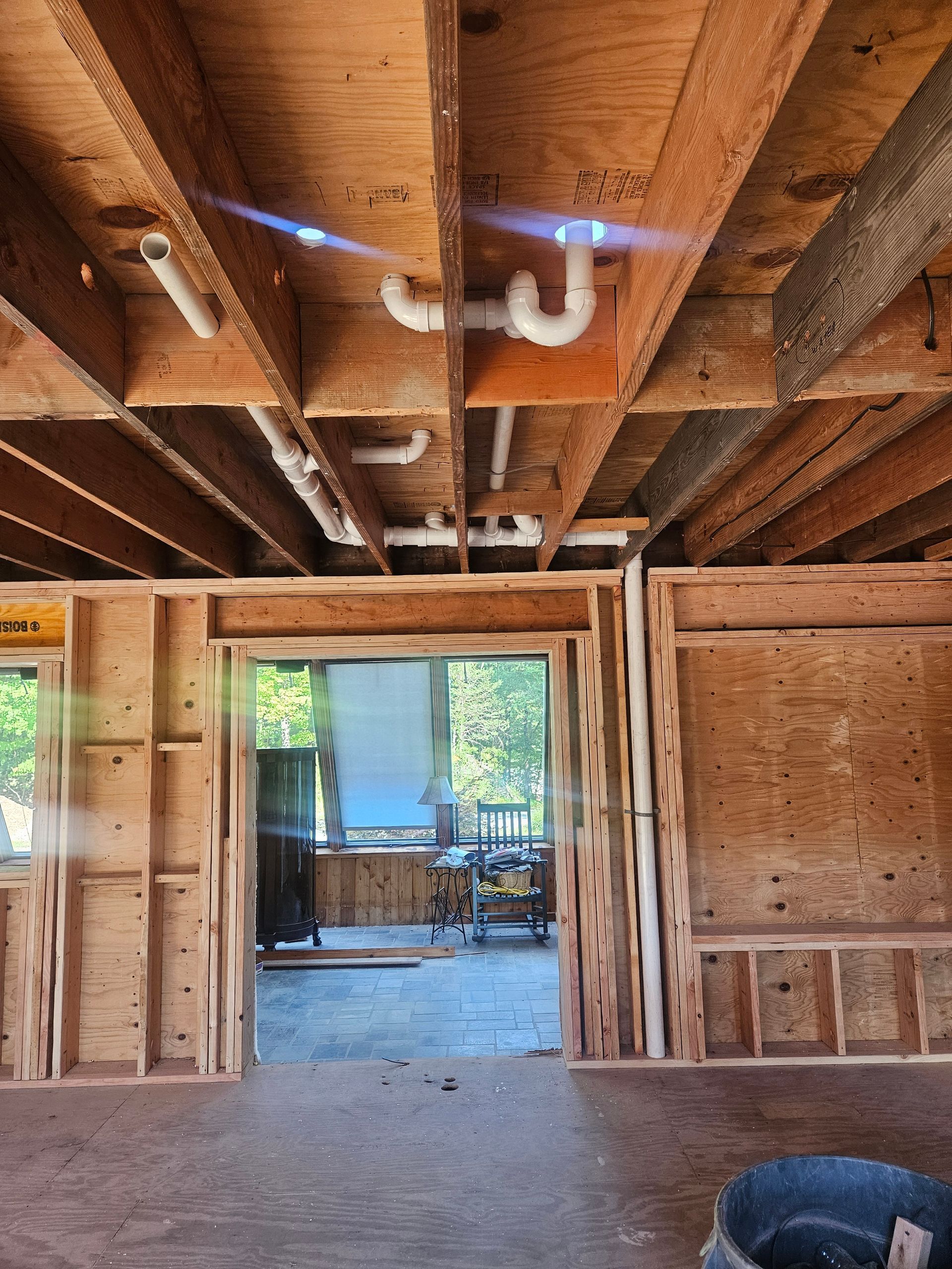 Framing of a room with exposed plumbing in the ceiling. Doorway leads to another room.