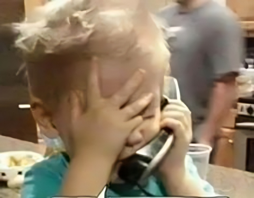 Blond toddler covers face while talking on a landline phone in a kitchen, adult in background.