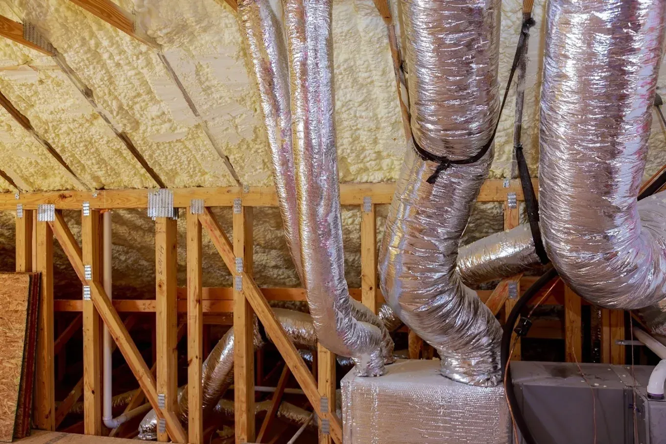 Attic with exposed wooden framing and insulated with spray foam. Metallic air ducts are visible.