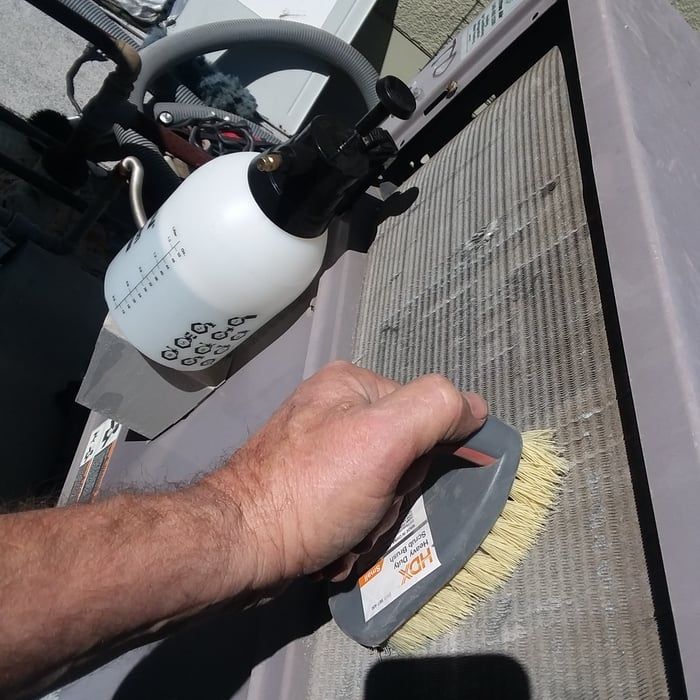 Person cleaning an AC unit with a spray bottle and brush, outdoors.