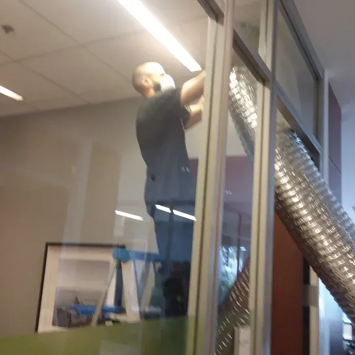 Man on a ladder installing something near a window. Fluorescent lights and a metal vent pipe are visible.