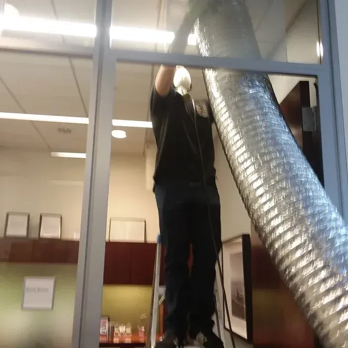 Person on a ladder installs silver ductwork near a window in an office setting.