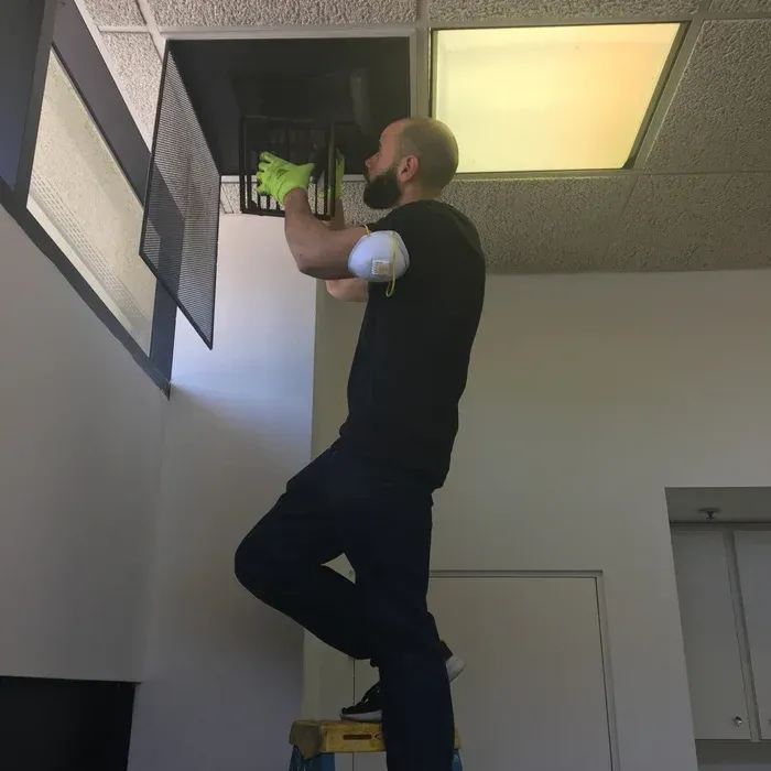 Man on a ladder cleaning a ceiling vent, wearing gloves and a face mask in an office.