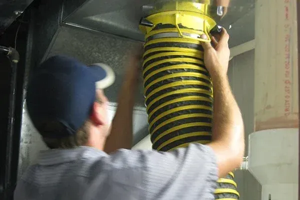 Person in blue cap attaching a flexible yellow and black air duct to a metal air vent.