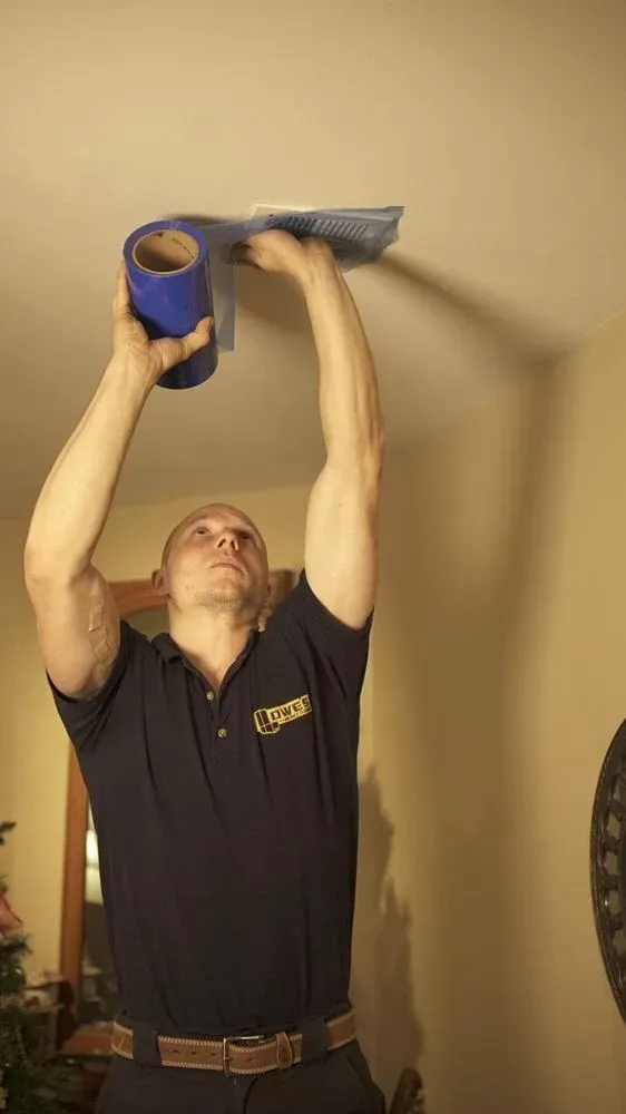 Man in blue shirt taping something on the ceiling. Blue tape roll in one hand, reaching upwards. Beige wall.