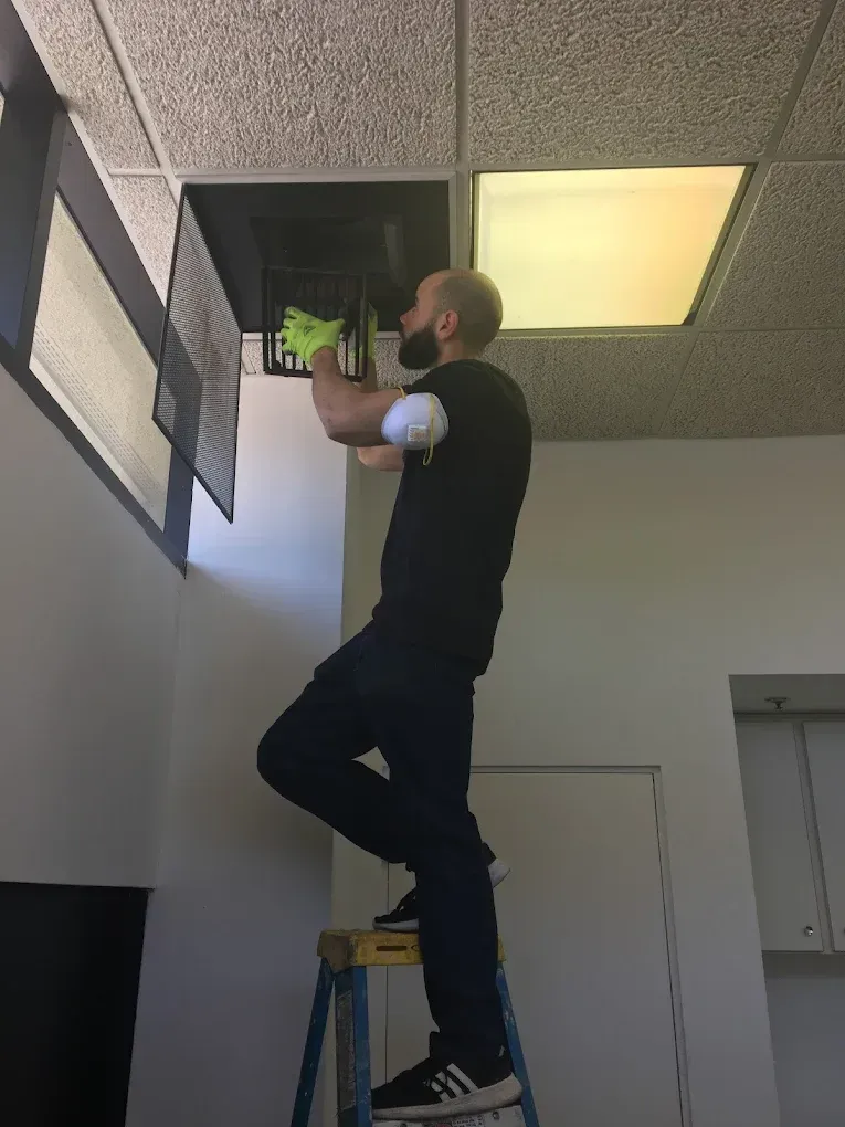 Man on a ladder cleaning a ceiling vent in an office.