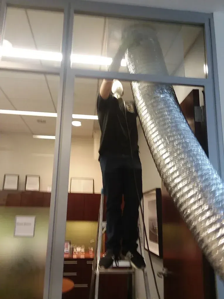 Person on a ladder installing a large, silver vent tube near a glass doorway.