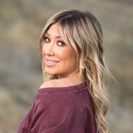 Woman with long brown hair, blue eyes, wearing a pink hoodie, smiling.