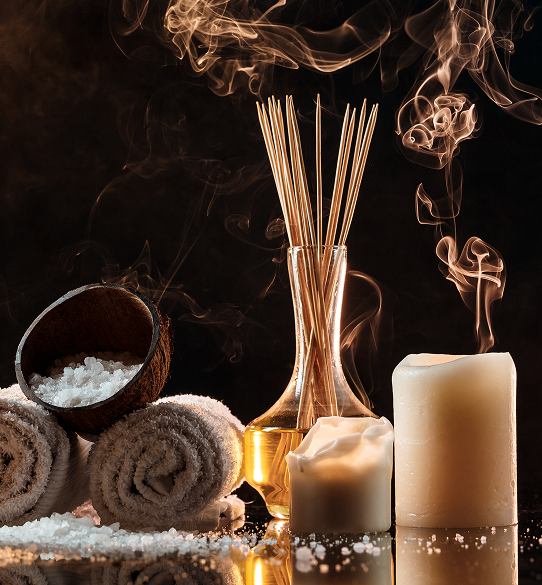 Spa scene: towels, bowl of salt, candles, reeds in oil, and smoke against a dark background.