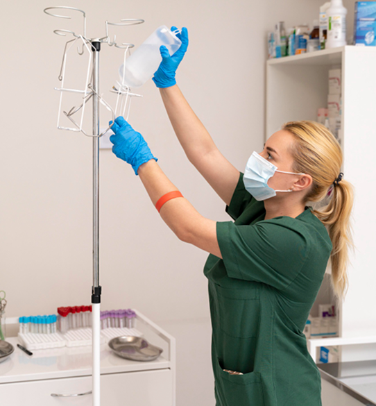 A healthcare worker in green scrubs hangs an IV bag on a stand in a medical setting.