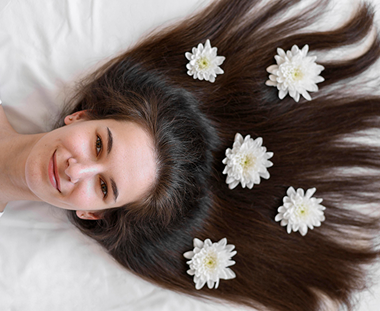 Woman smiling, lying on white sheet, dark hair adorned with white flowers.