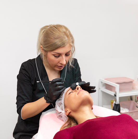 Woman performing eyebrow microblading on another woman in a salon.