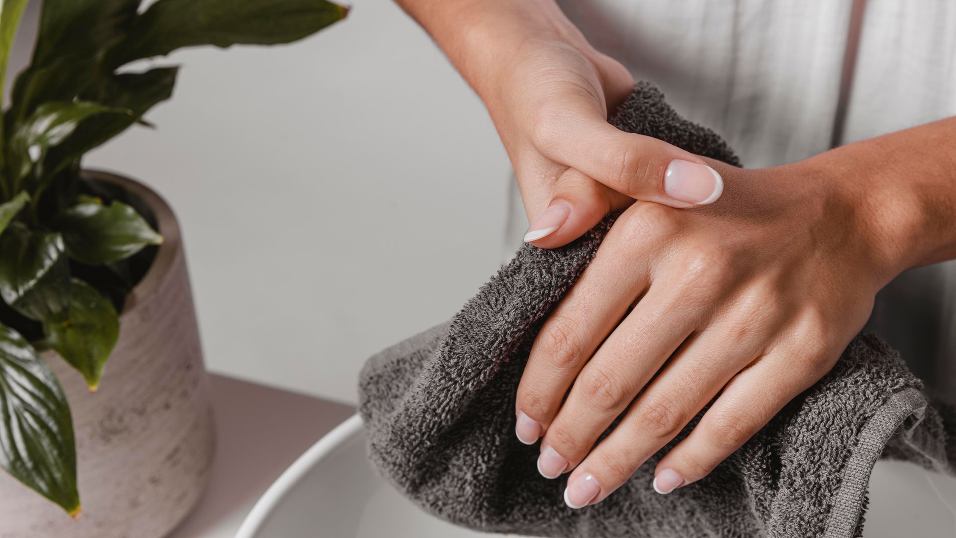 Woman drying hands with a gray towel next to a sink and a potted plant.