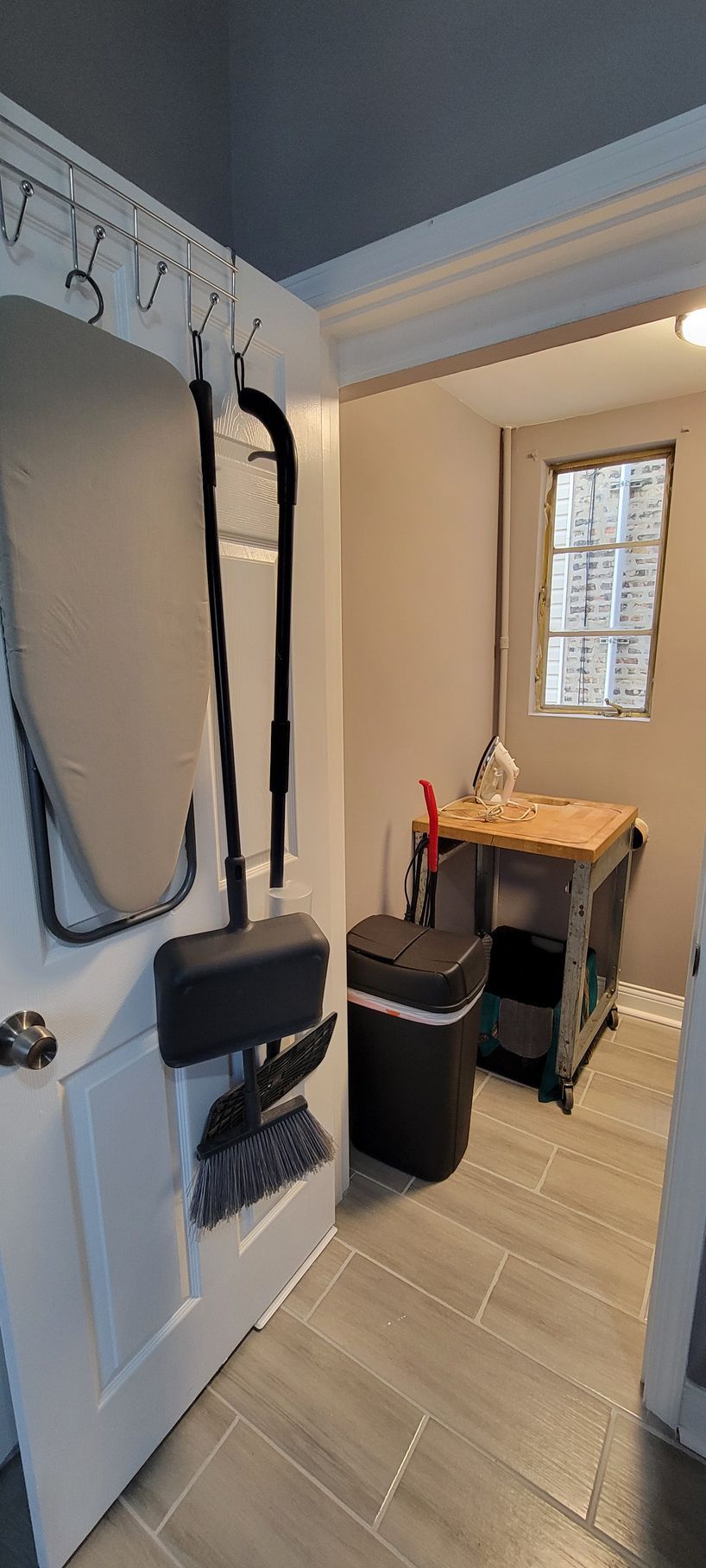 A laundry room with an ironing board and a trash can.