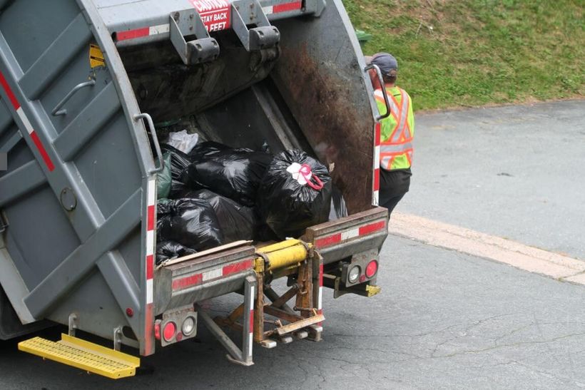 Junk Removal contractor riding on the side of a dumpster truck filed with black trash bags.