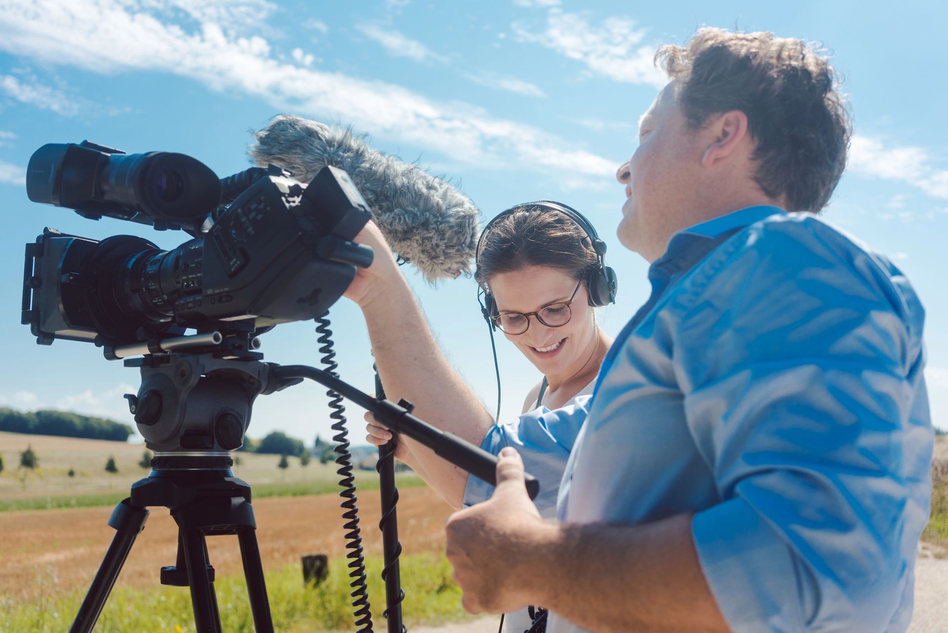 Dos personas filmando al aire libre con una cámara profesional y un micrófono, cielo azul y campo de fondo.