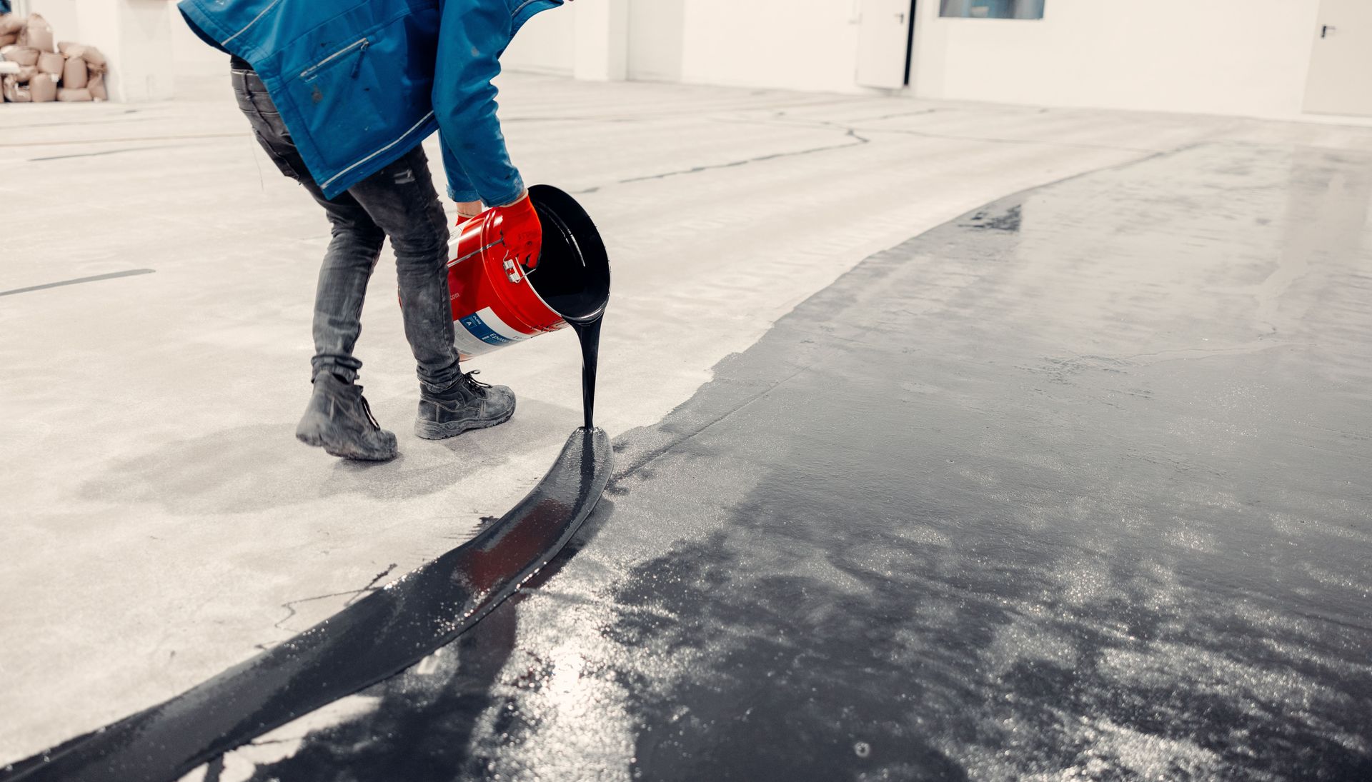 Person pouring black sealant from a bucket onto a concrete floor.