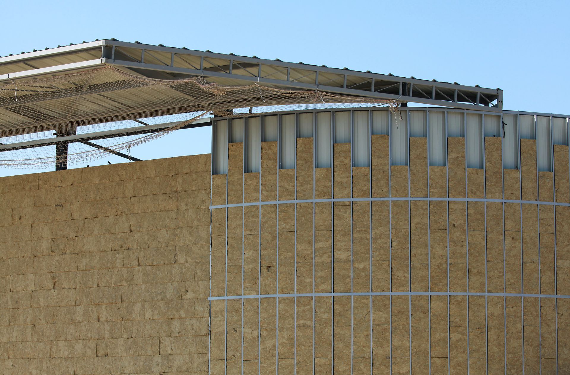 Circular building with tan walls and metal support structure, under a shaded roof.