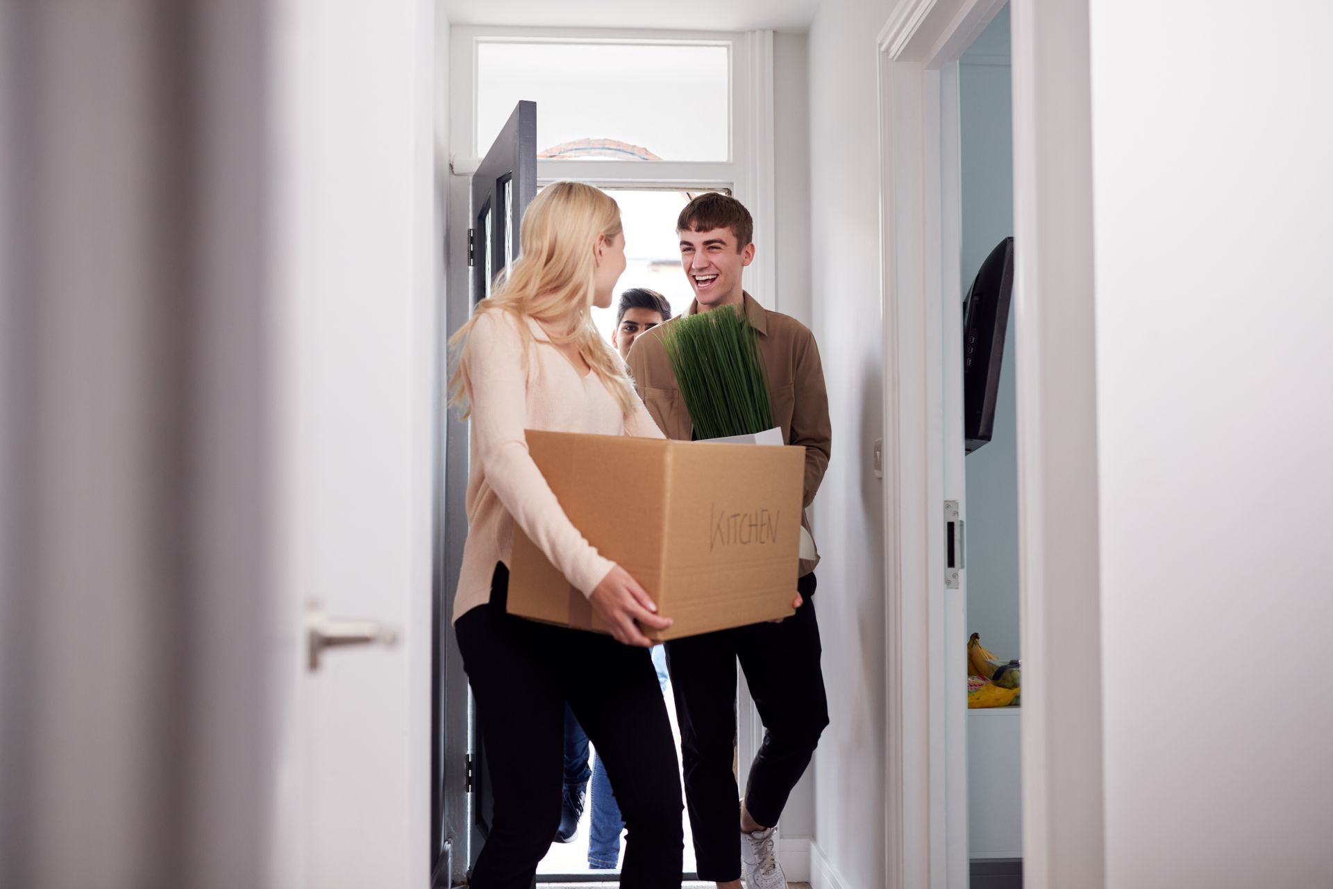 Woman and man carrying boxes and a plant into a brightly lit hallway. A third person stands in the background.