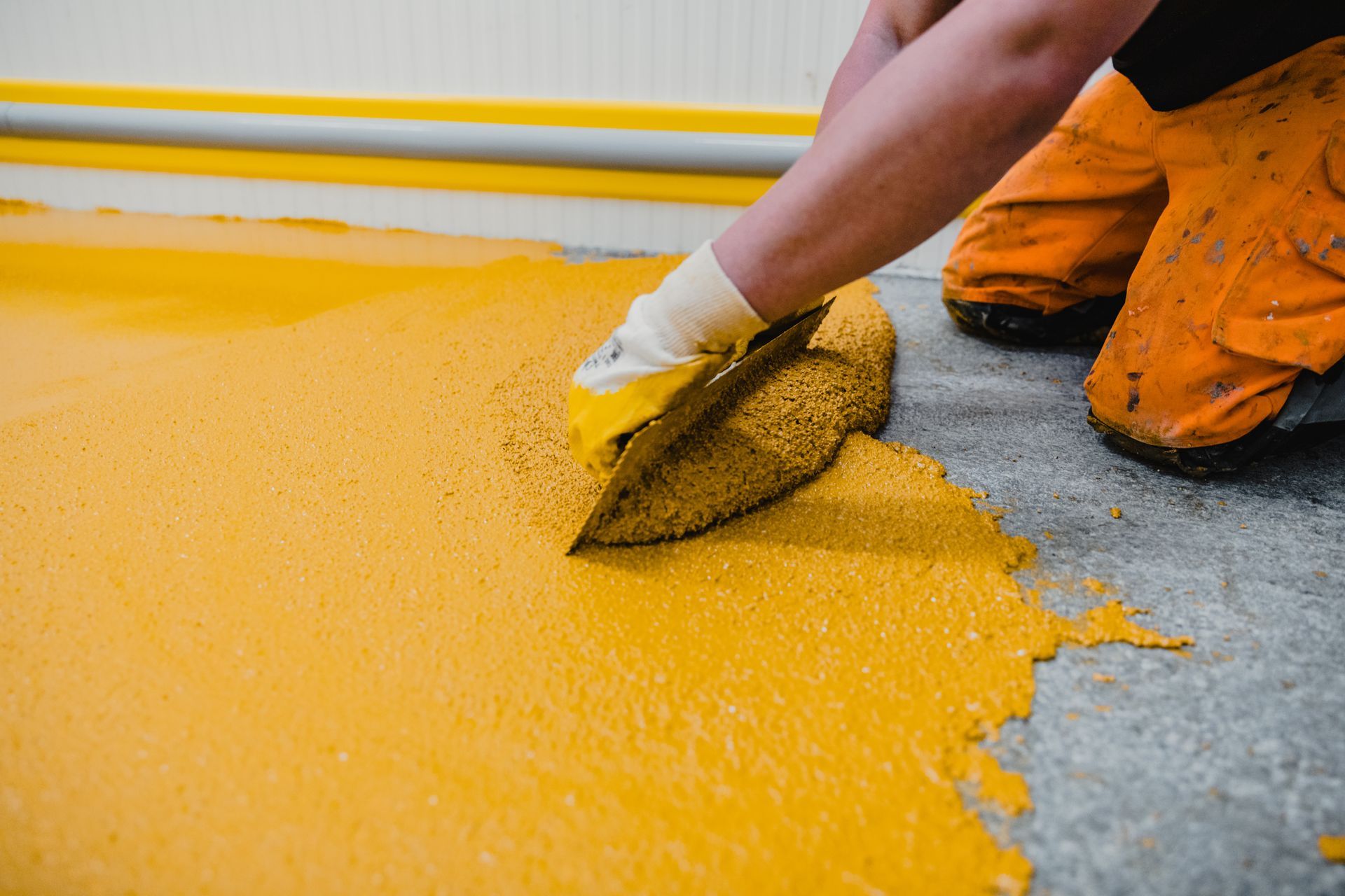 Person smoothing yellow epoxy flooring with a trowel, wearing gloves and orange work pants.