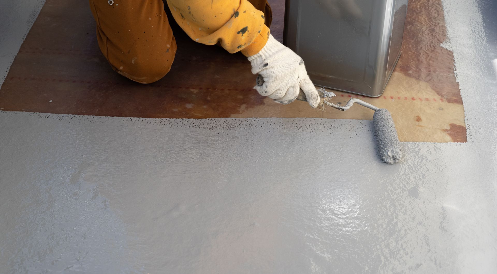 Person in gloves paints a floor with a roller and gray paint.