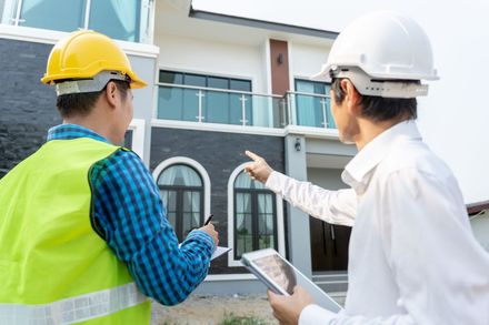 Two construction workers are looking at a tablet in front of a house.