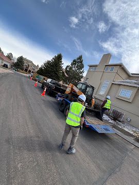 Roadwork crew loading equipment onto a flatbed truck on a residential street, with traffic cones and workers in safety vests.