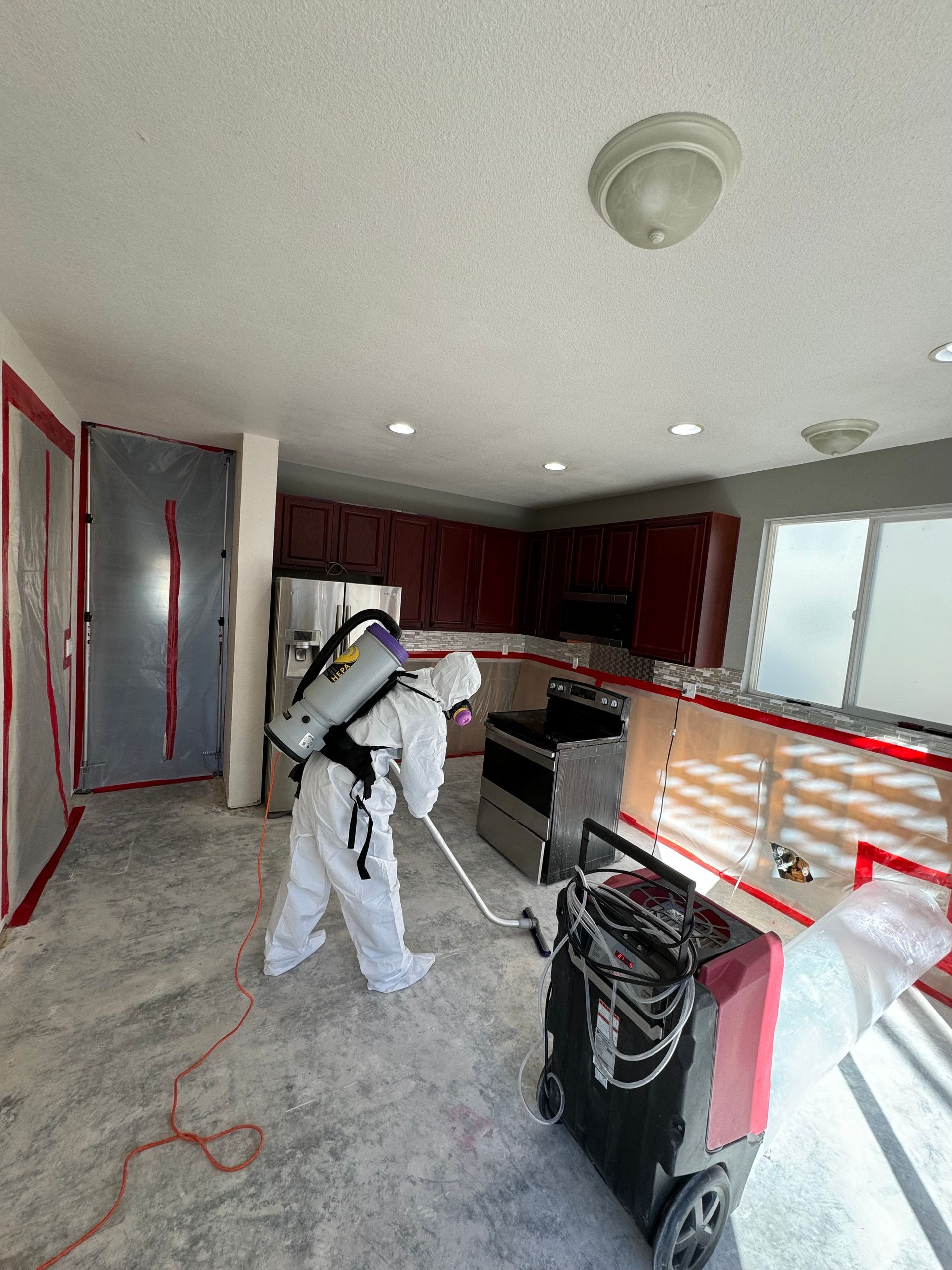 Worker in white protective suit spray painting a kitchen interior with cabinets and counters.