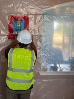 Worker in hard hat and reflective vest installing a red warning sign on plastic-covered wall.