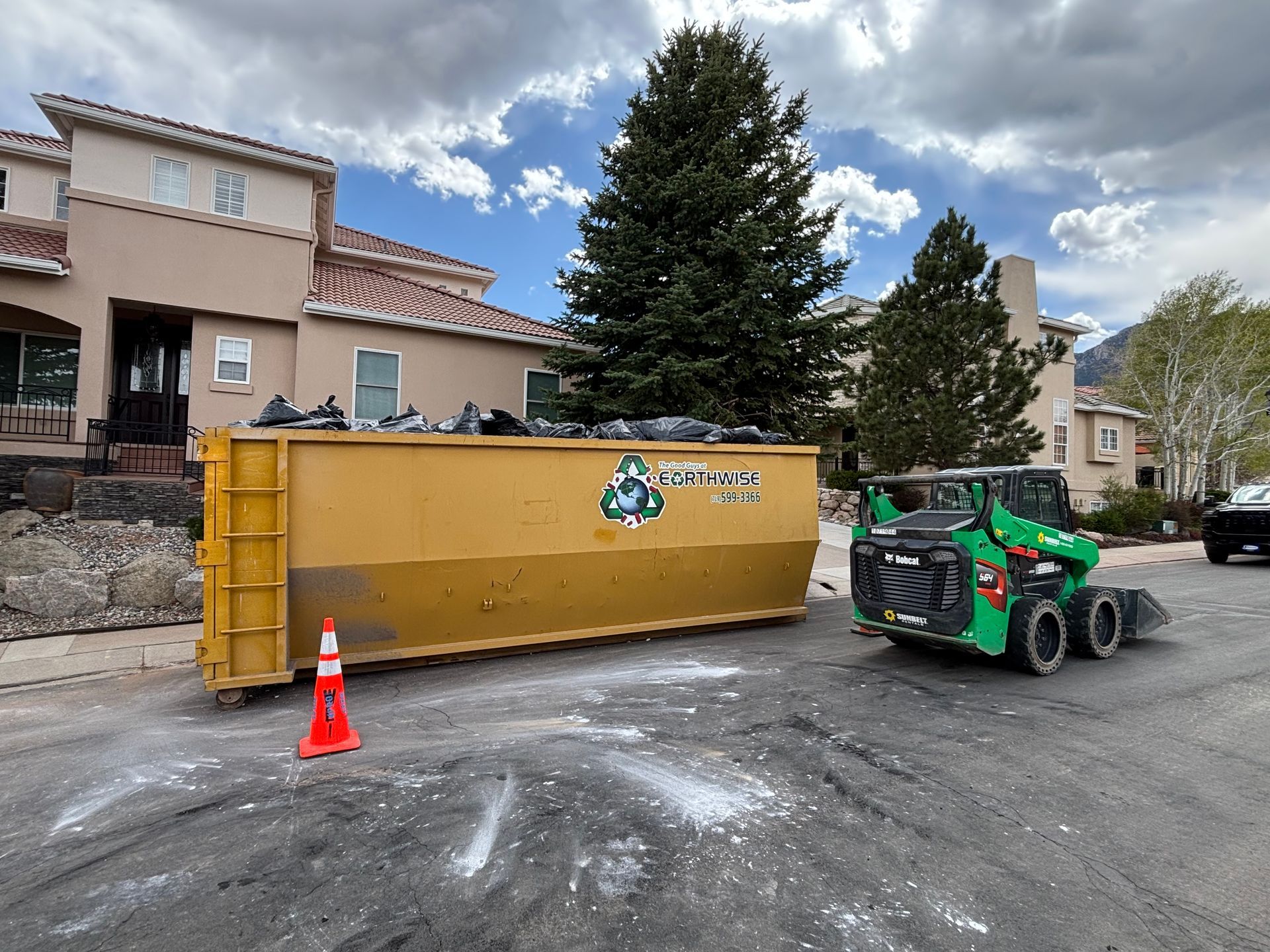 Yellow dumpster beside a green skid steer in a parking lot outside an apartment building, with an orange cone nearby.