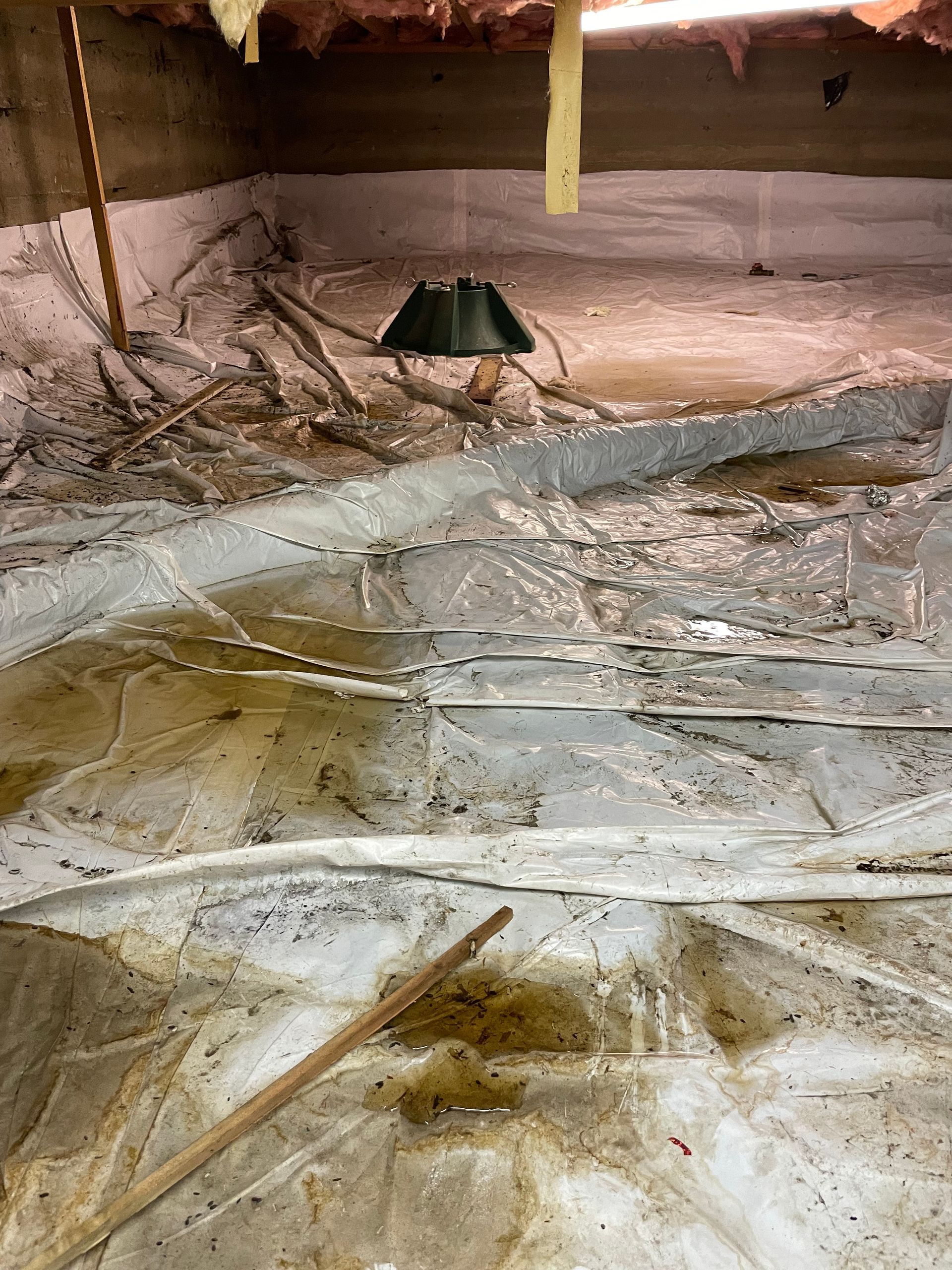 Crawlspace with torn plastic sheeting, debris, and a small black device on the dirt floor
