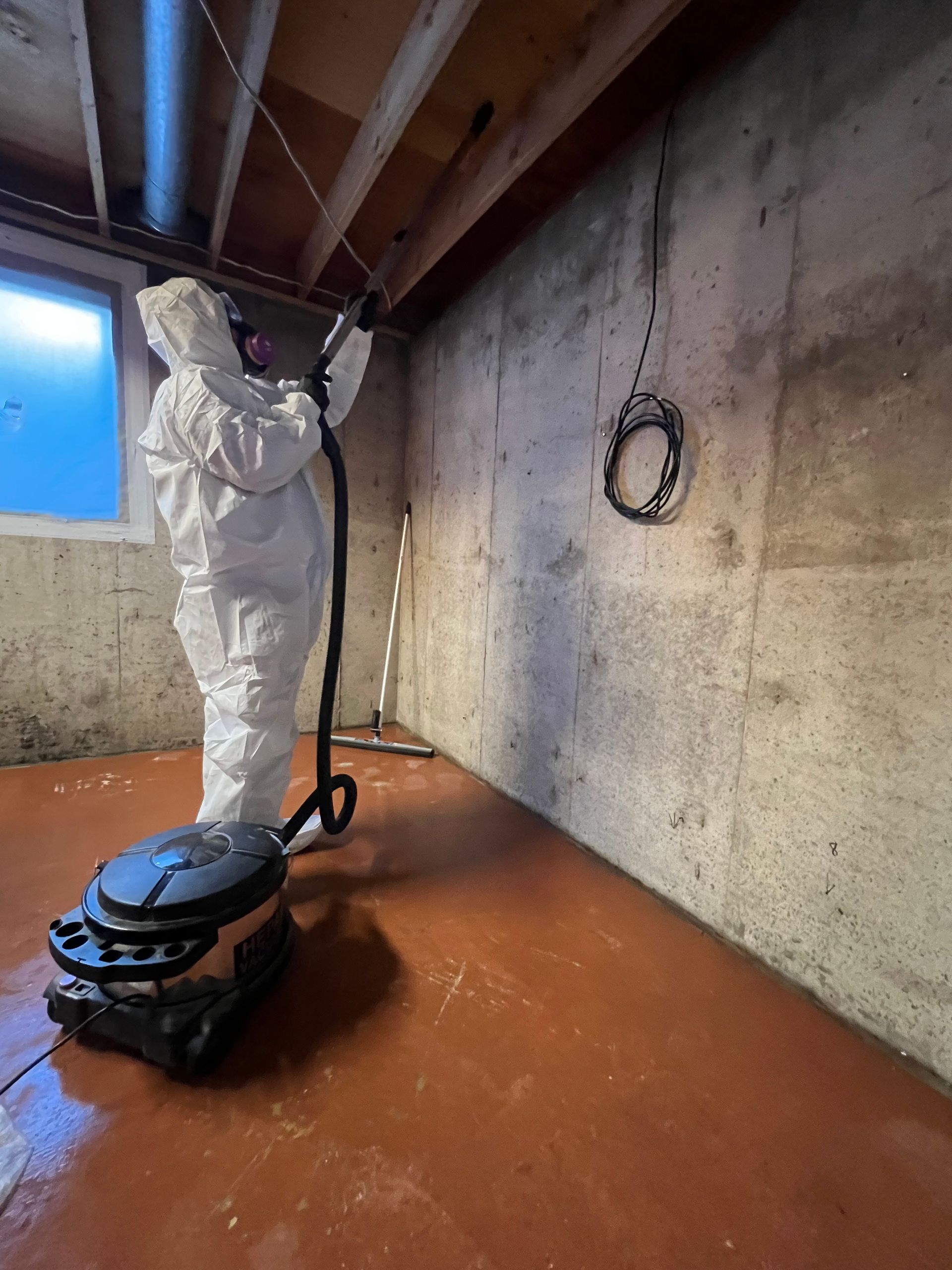 Worker in white protective suit painting a basement ceiling, with a black paint tray on the floor.