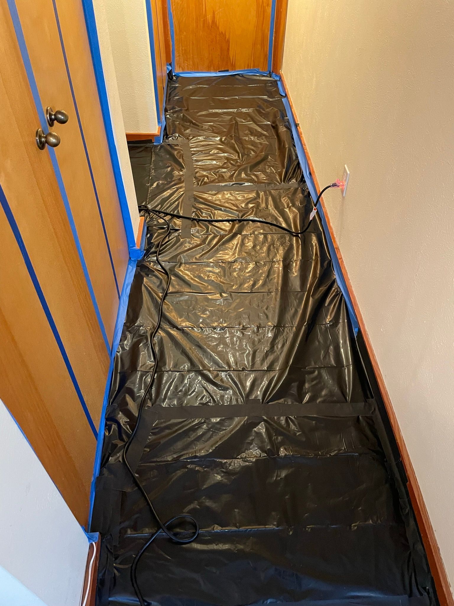 Narrow hallway covered with black plastic sheeting, with wooden closet doors on the left and a bare wall on the right