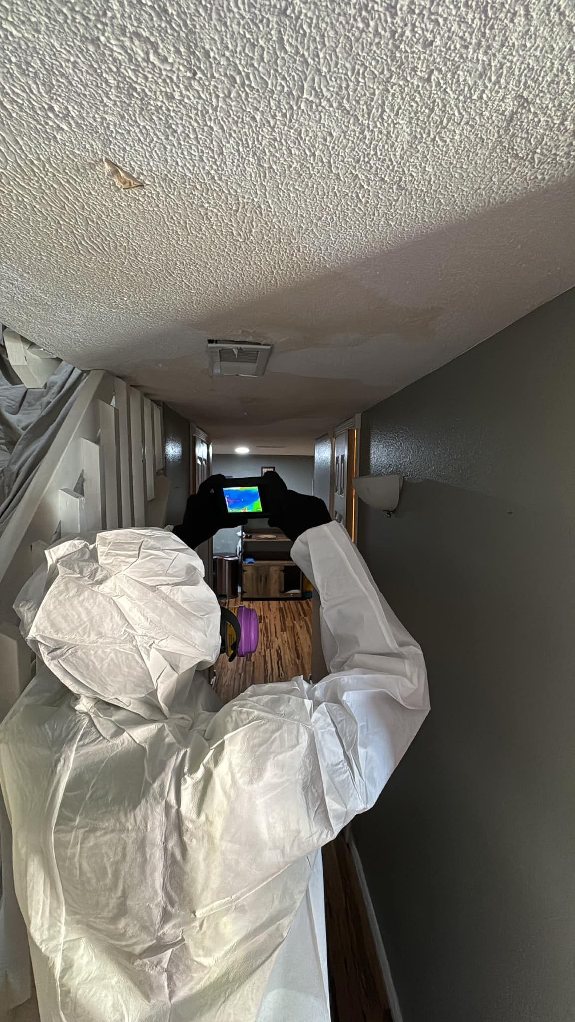 Person in white protective suit cleaning a narrow hallway with plastic-covered furniture and debris on the floor