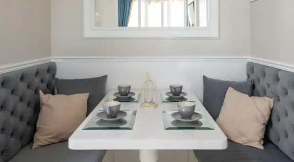 Ein weißer Esstisch mit vier Gedecken zwischen zwei grauen, gesteppten Sitzbänken mit beigefarbenen Zierkissen.